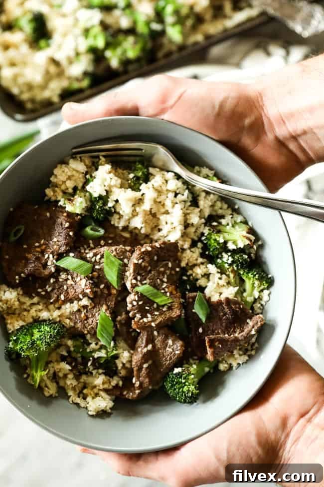 A bowl holding cauliflower rice, roasted broccoli, and sesame beef, garnished with chopped green onion and sesame seeds. 