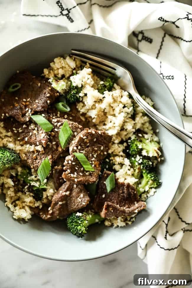 A bowl of cauliflower rice, broccoli, and sesame beef with a fork, garnished with green onions and sesame seeds. 