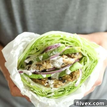 A hand holding a loaded turkey burger with a crisp lettuce bun and parchment paper wrapped around it, ready to eat.