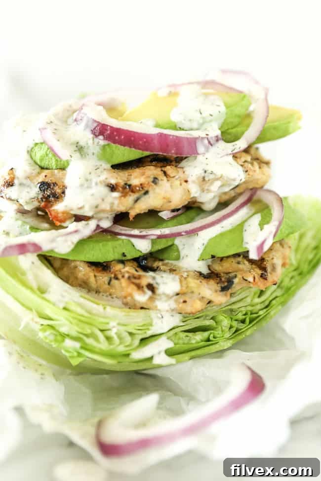 Close-up shot of a stack of Greek turkey burgers, artfully arranged on lettuce leaves and topped with fresh avocado slices, thinly cut red onion, and a delicate drizzle of creamy tzatziki sauce. The vibrant layers invite a delicious bite.