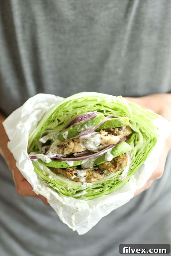 A hand holding a perfectly assembled Greek turkey burger, wrapped in a crisp lettuce bun and parchment paper, showcasing the fresh toppings and the delicious patty.