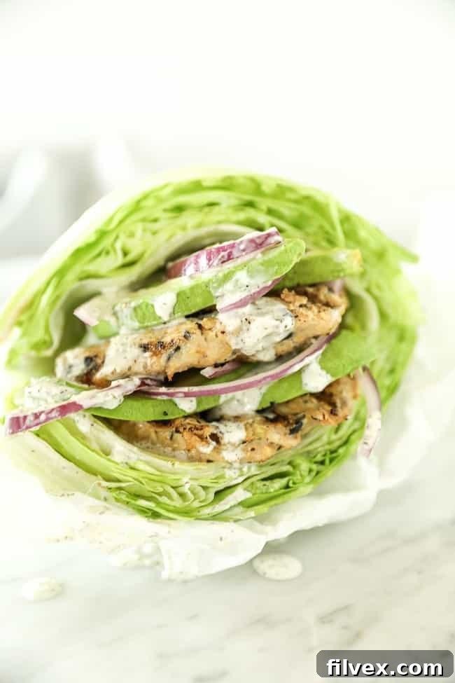 Two fully assembled Greek turkey burgers, stacked high between crisp lettuce buns. They are generously topped with sliced avocado and red onion, with the dairy-free tzatziki sauce artfully drizzled over, creating an inviting presentation.