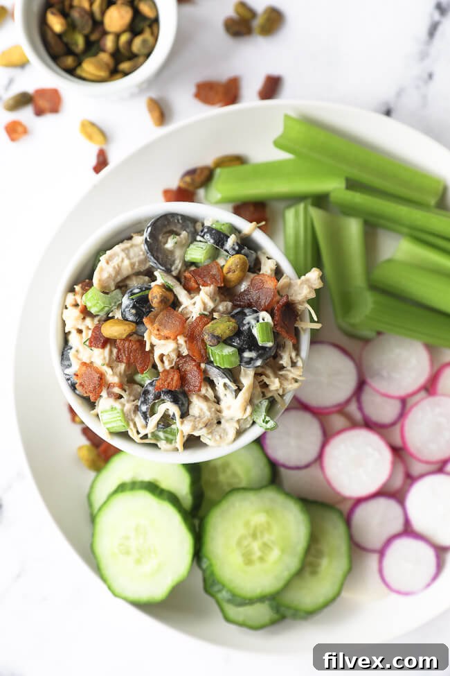 Vertical overhead image of chicken salad in a bowl on a plate with cucumber slices, radish slices and celery sticks, illustrating serving suggestions.