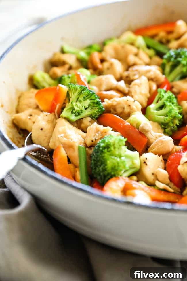 Angled close up image of cashew chicken in a skillet with a serving spoon. Chicken, cashews, broccoli, red bell pepper and an asian sauce.