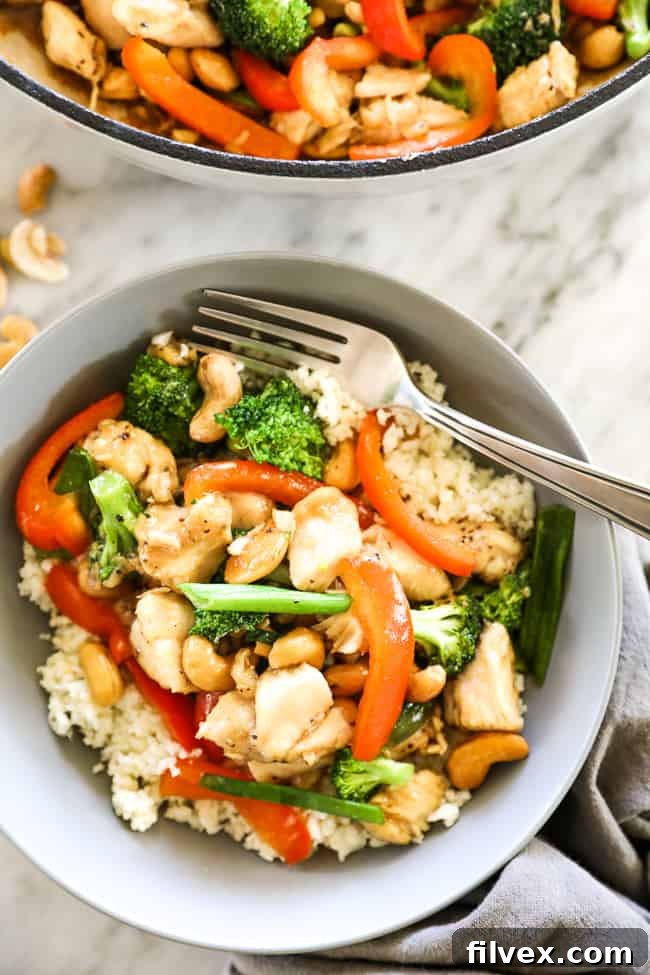 Overhead close up image of cashew chicken served in a bowl over cauliflower rice with a fork. Chicken, cashews, broccoli, red bell pepper and an asian sauce.