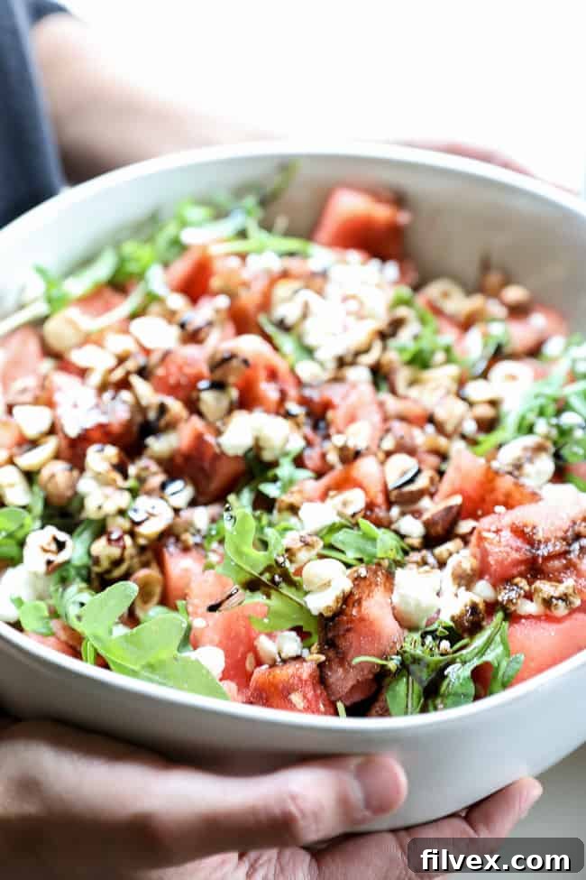 Angled vertical image of holding a large serving bowl of refreshing watermelon salad with arugula, hazelnuts, balsamic glaze, and optional feta cheese.