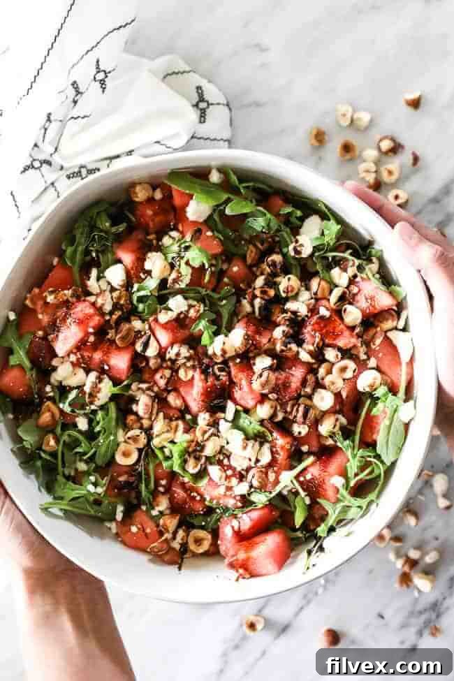 Overhead vertical image of a large serving bowl filled with a refreshing watermelon salad, featuring cubed watermelon, fresh arugula, chopped hazelnuts, a balsamic drizzle, and optional crumbled feta cheese.