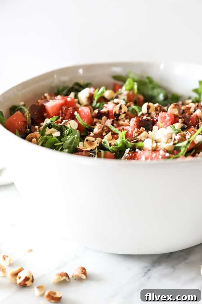 Angled vertical image of a large serving bowl of vibrant watermelon salad, showcasing the fresh watermelon, peppery arugula, crunchy hazelnuts, balsamic vinegar, and optional feta cheese.