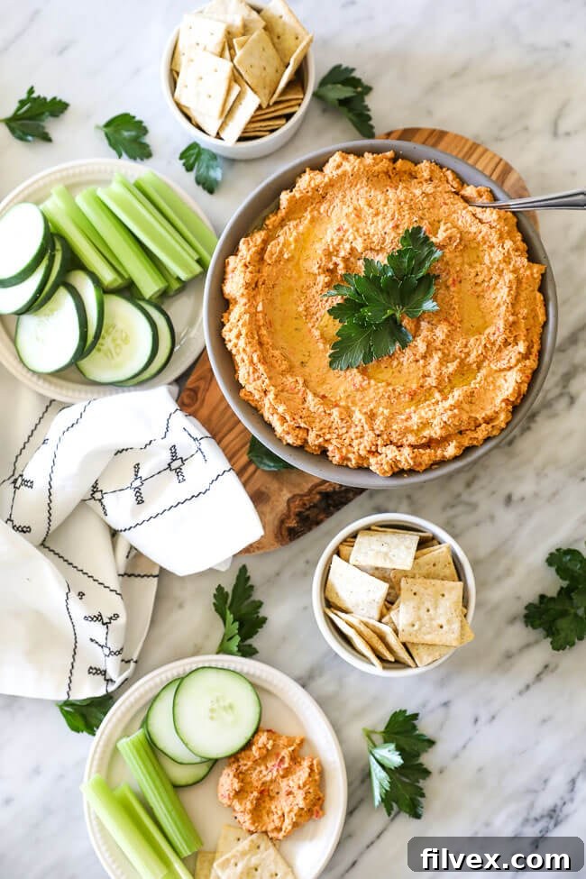 A beautifully presented bowl of easy cauliflower hummus, surrounded by an array of colorful fresh vegetables and grain-free crackers, all spread out on a white marble surface. The image is a vertical shot highlighting the spread.