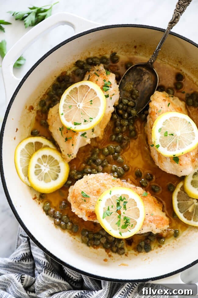 Vertical overhead image of healthy Lemon Chicken Piccata in a large skillet with a bright lemon-caper sauce, fresh lemon rounds, and chopped parsley. A serving spoon rests in the skillet, ready for a delicious meal. This image showcases a vibrant and inviting weeknight dinner.
