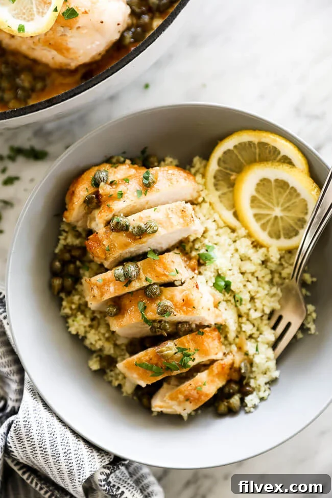 Vertical overhead close-up image of healthy Lemon Chicken Piccata served in a bowl over fluffy cauliflower rice, adorned with bright lemon rounds and a sprinkle of fresh chopped parsley. A fork is dug into the bowl, highlighting the tender chicken and rich sauce. This enticing shot emphasizes the dish's health and flavor.