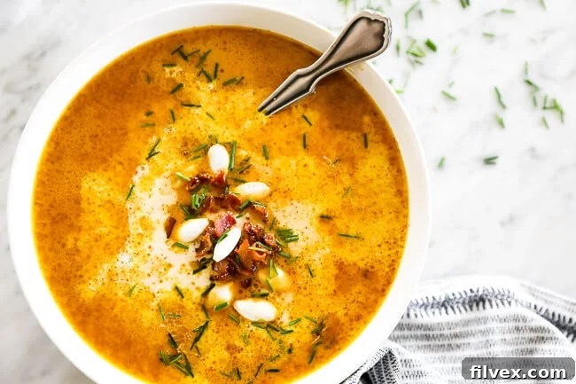 Horizontal overhead image of pumpkin soup in a bowl with a spoon. Topped with bacon bits, pumpkin seeds and chopped chives. 