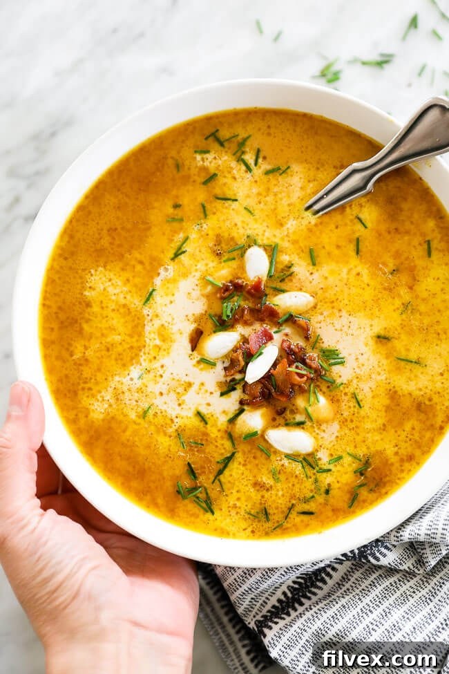 Vertical overhead image of holding a bowl of pumpkin soup with a spoon. Topped with bacon bits, pumpkin seeds and chopped chives. 
