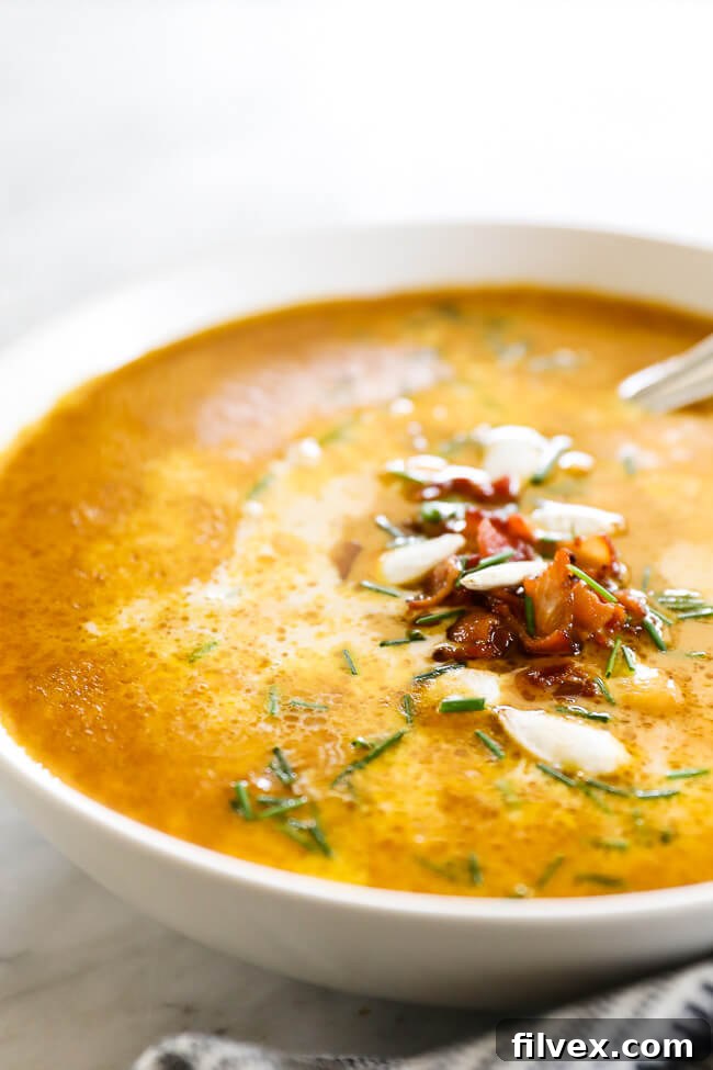 Vertical angled image of pumpkin soup in a bowl. Topped with bacon bits, pumpkin seeds and chopped chives. 