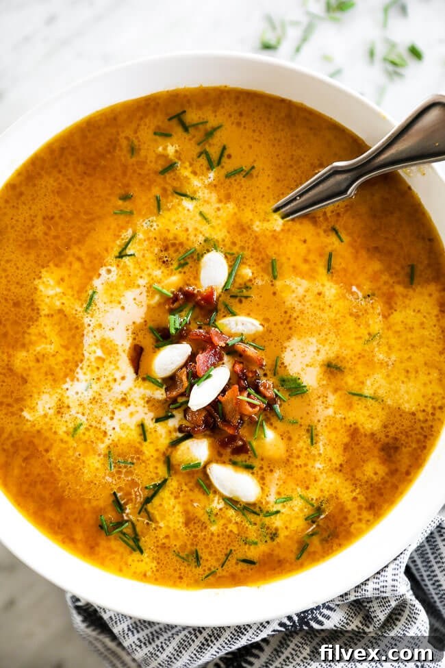 Vertical overhead close up image of pumpkin soup in a bowl with a spoon. Topped with bacon bits, pumpkin seeds and chopped chives. 