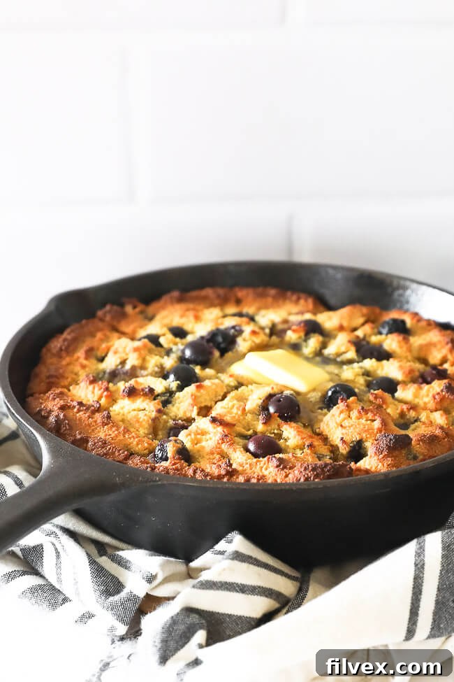 Vertical angled shot of blueberry cornbread in a cast iron skillet. Cut up into pieces with grass fed butter melting on top. 