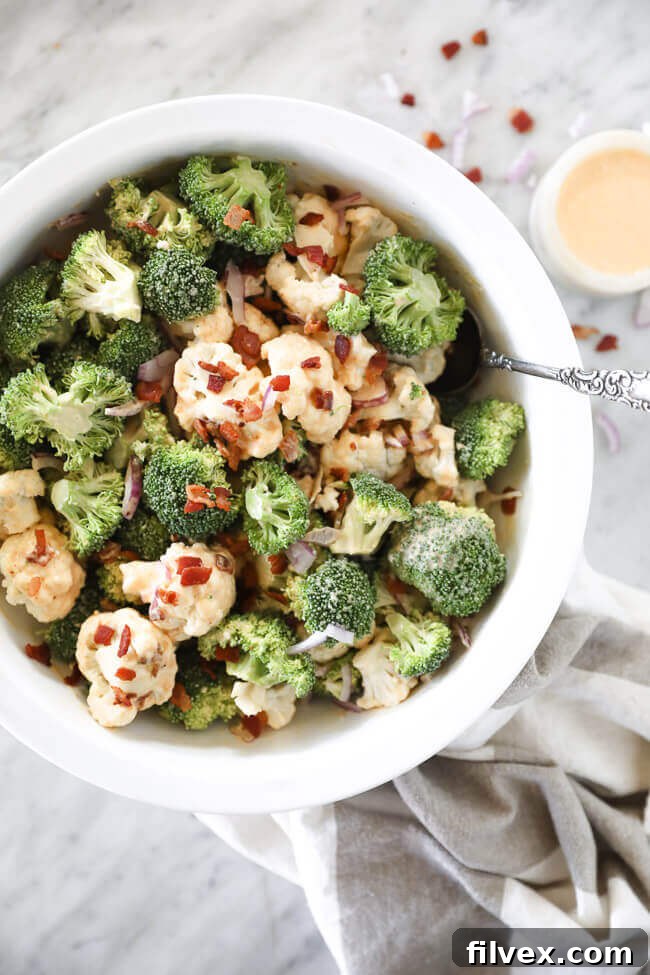 A vibrant bowl of creamy keto broccoli and cauliflower salad with crispy bacon bits and diced red onion. The dressing is drizzled generously over the fresh florets.