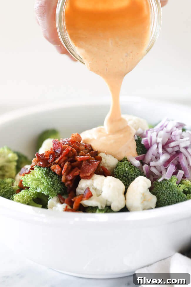Close-up shot of crisp broccoli and cauliflower florets mixed with red onion and crumbled bacon, with creamy aioli dressing being poured from above, highlighting the fresh ingredients.