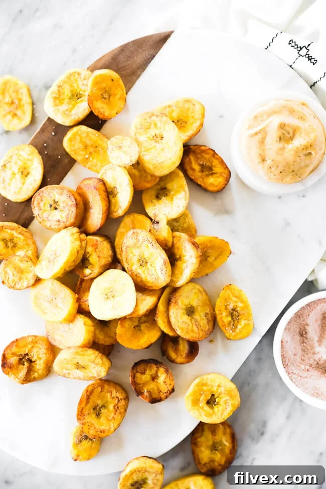 Vertical overhead image of pan fried plantains, some piled and some scattered on a stylish marble board, accompanied by a small bowl of dipping sauce and a sprinkle of sea salt.