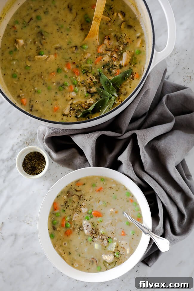 Vertical image of chicken pot pie soup served in a bowl with a spoon in it. Large soup pot in the image as well with ground pepper on the side. 