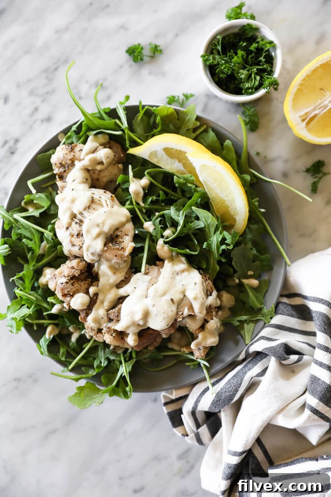 Chicken thighs covered in creamy sauce on top of arugula on a plate with lemon wedges vertical overhead image