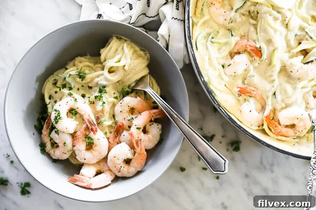 Horizontal overhead image of creamy garlic shrimp with zoodles served in a bowl with a fork with noodles wrapped around it. Skillet of dish off to the side and parsley sprinkled on top. 