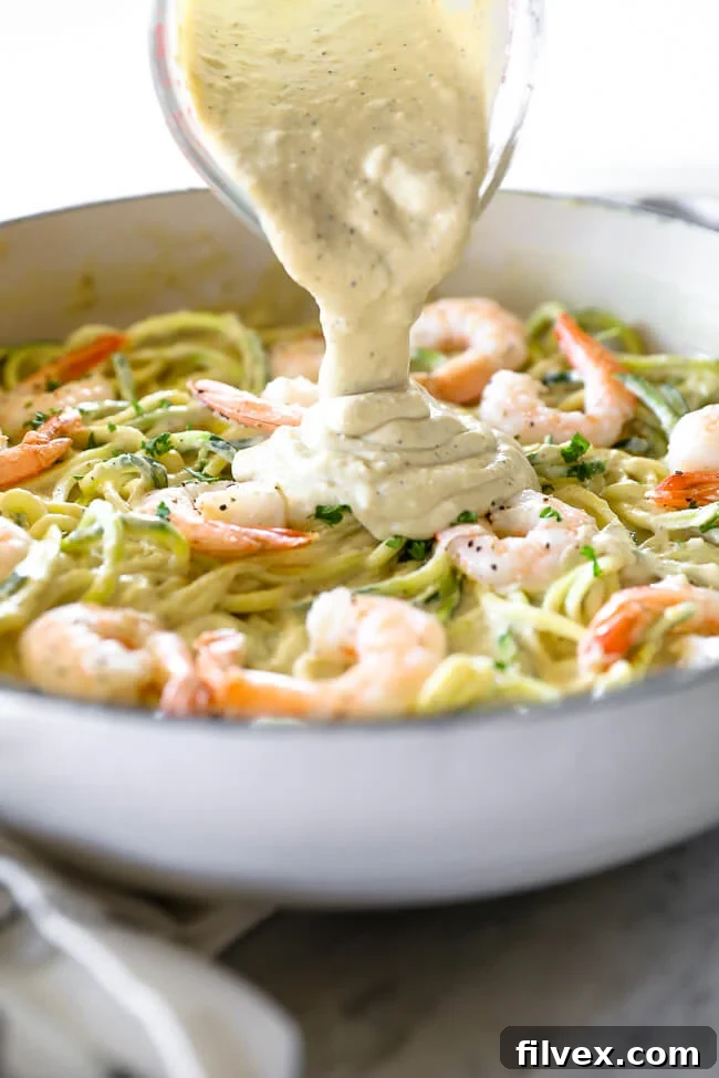 Angled image of pouring creamy sauce over skillet of zucchini noodles and shrimp. 
