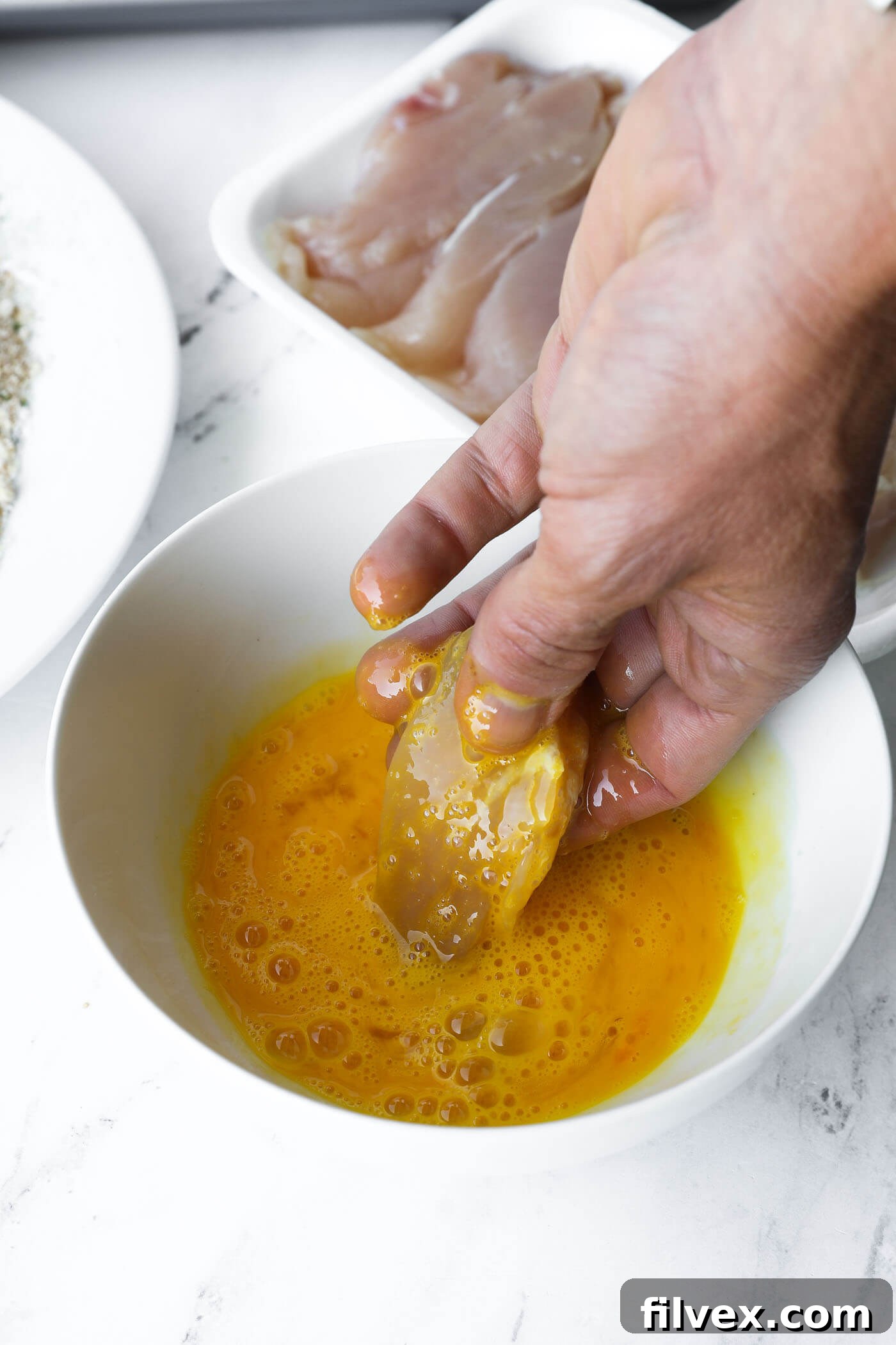 A chicken tender being coated in whisked egg using the 'wet hand' technique.