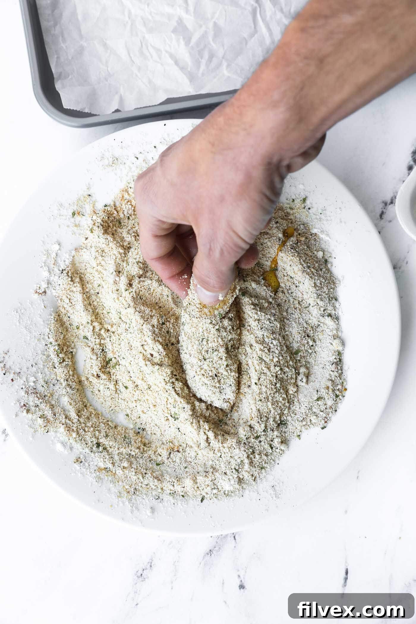 A chicken tender being dredged in the almond flour mixture using the 'dry hand' technique.