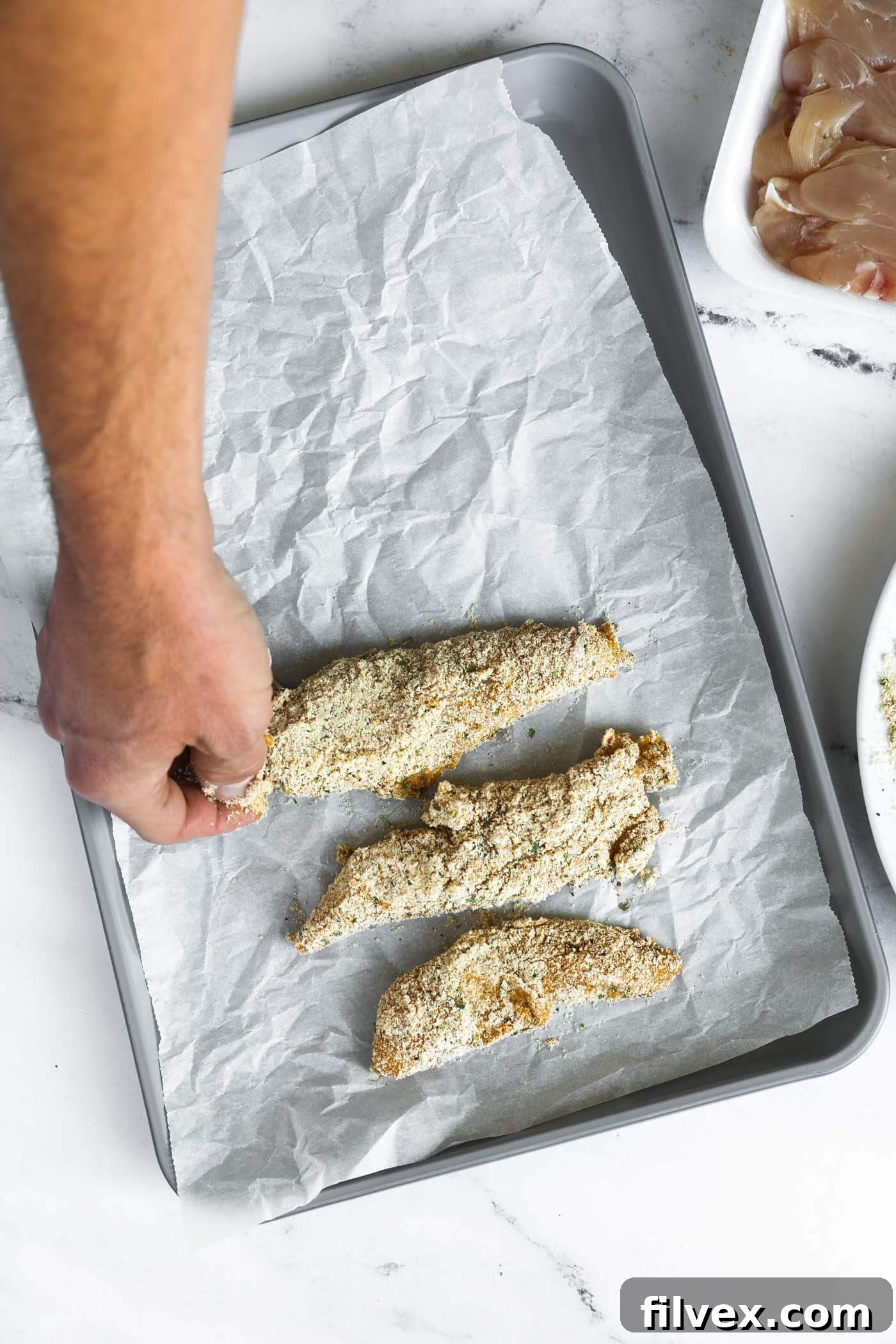 Multiple chicken tenders coated and arranged on a baking sheet.