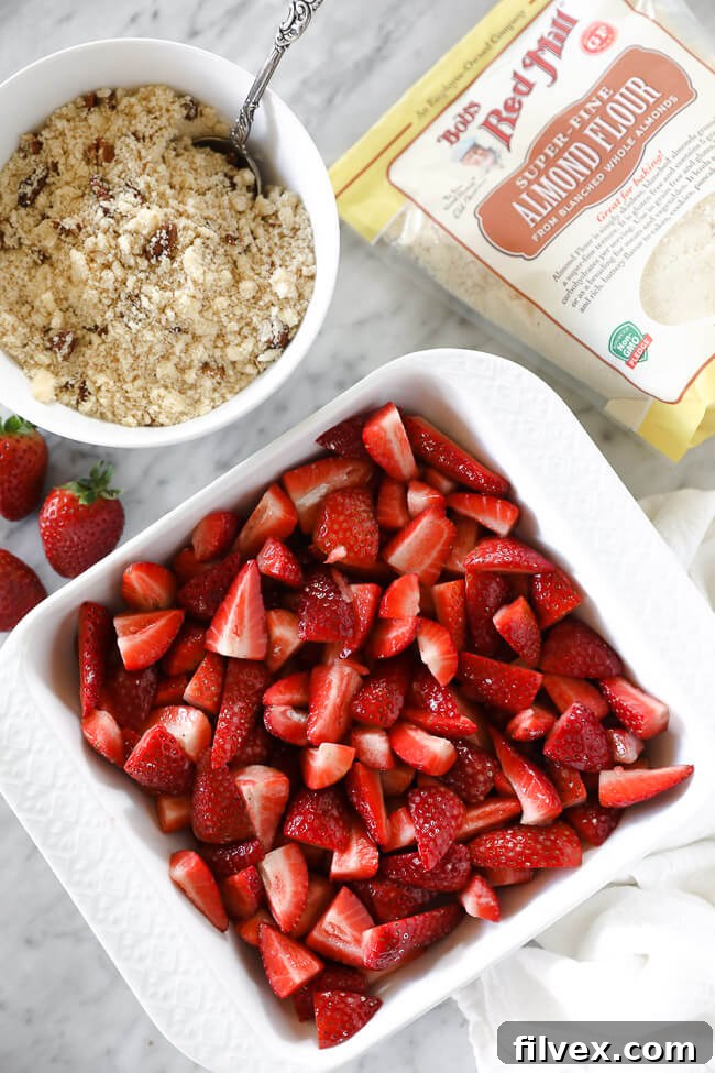 Vertical overhead image of strawberries in a baking dish with topping in a small bowl on the side and bob's red mill almond flour.