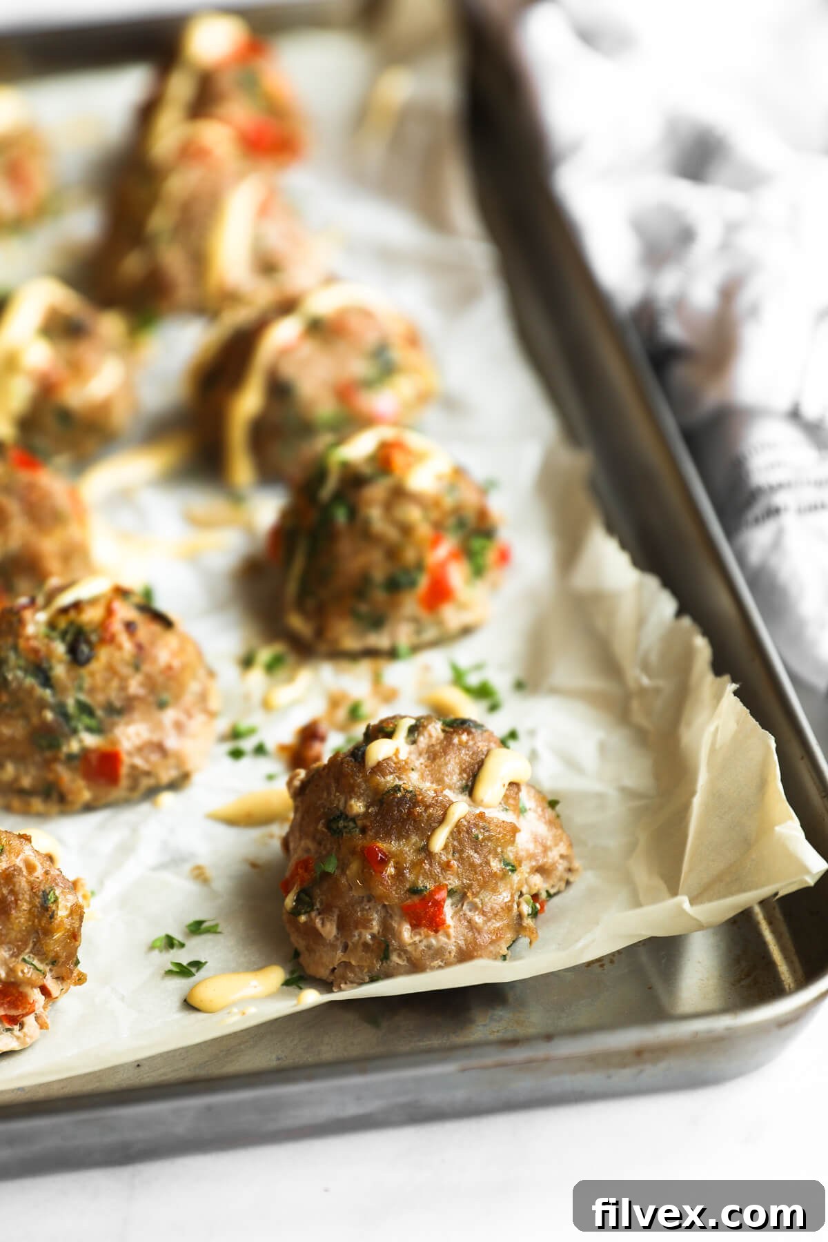 Angled close-up shot of perfectly golden-brown turkey meatballs on a baking sheet, with a rich, spicy aioli sauce artfully drizzled over them. The foreground meatball is in sharp focus, highlighting its texture.