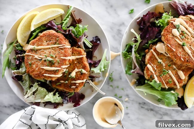 Horizontal overhead image of salmon patties on a plate served over fresh greens and topped with drizzle of spicy aioli sauce. 