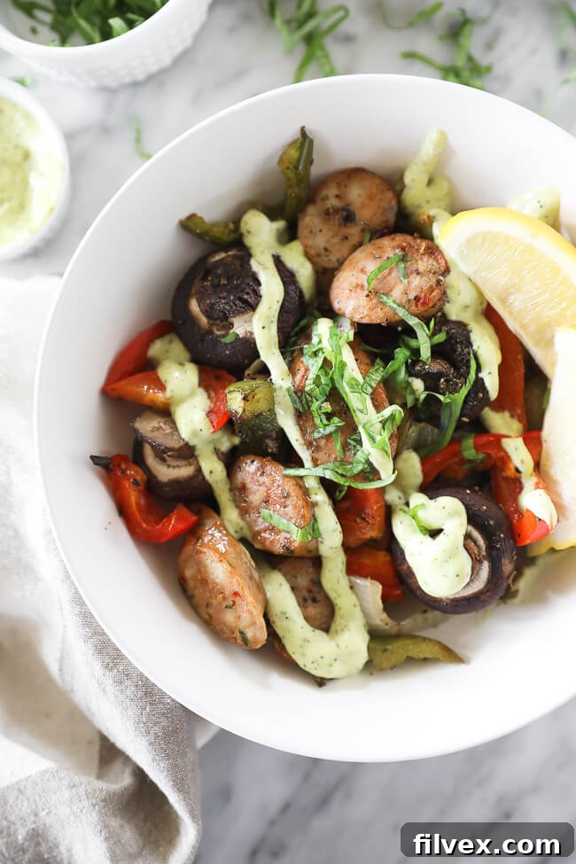 Vertical overhead image of a serving bowl featuring Italian sausage and peppers, generously topped with a vibrant herb aioli and freshly chopped basil. A sheet pan with more roasted ingredients is visible in the soft-focus background.