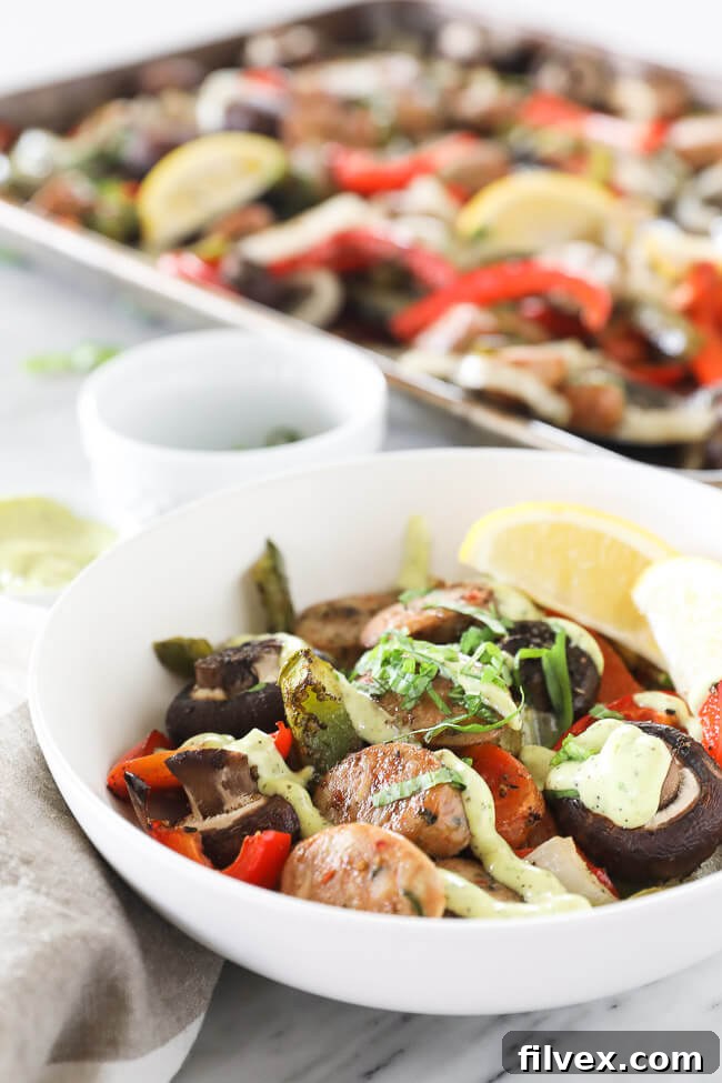 Vertical angled shot of a serving bowl featuring the Italian sausage and peppers recipe, beautifully plated with a generous drizzle of homemade herb aioli on top. The sheet pan with more roasted ingredients is visible in the background, hinting at more deliciousness.