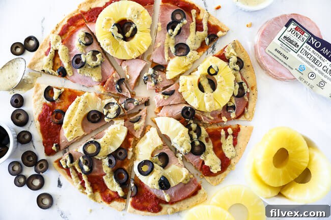 Horizontal image of hawaiian pizza cut into slices with olives, pineapple, canadian bacon, marinara sauce and a creamy dairy free sauce drizzled on top.