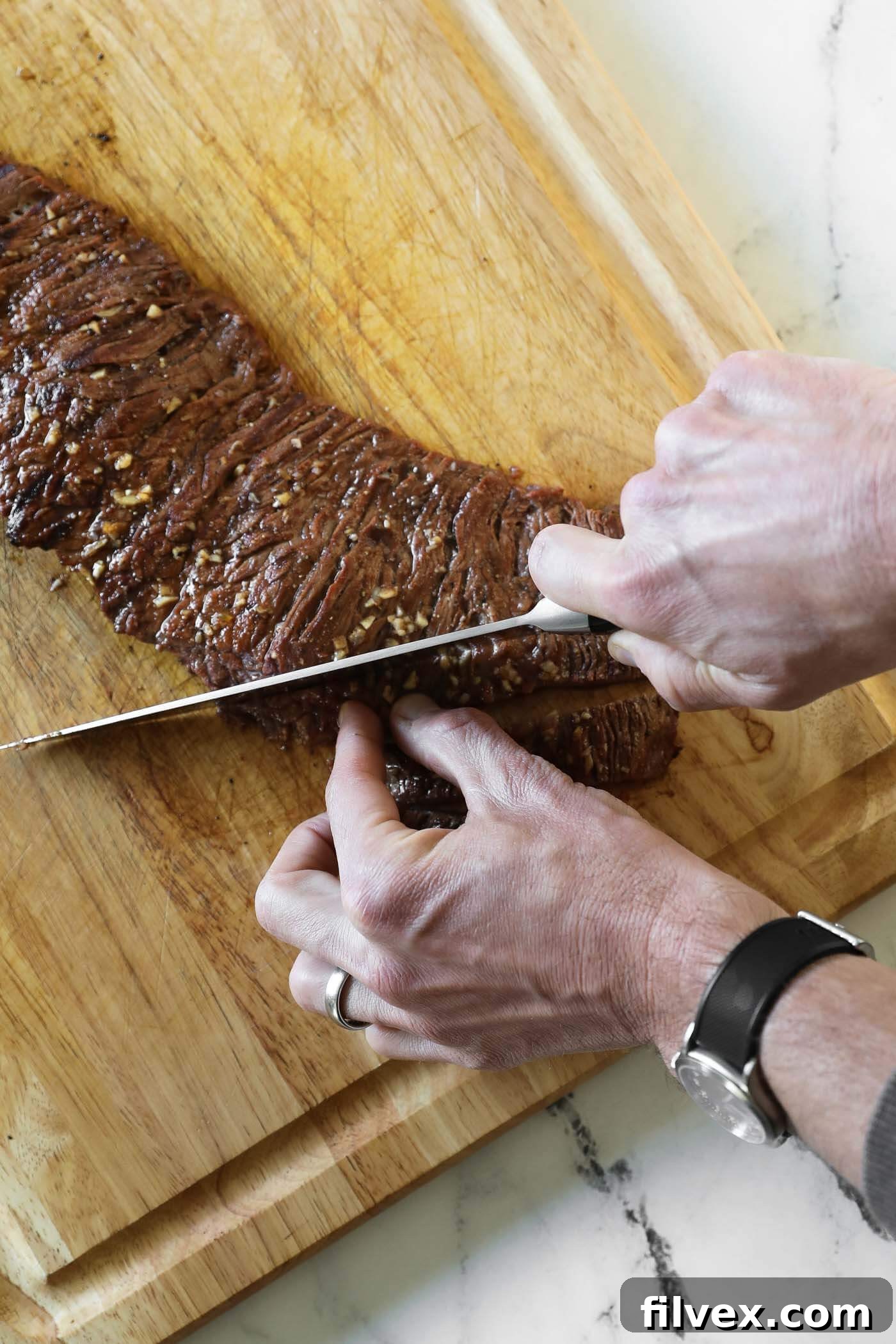 Overhead shot of hands and a knife carefully cutting a rested carne asada steak into thin strips against the grain on a wooden cutting board.