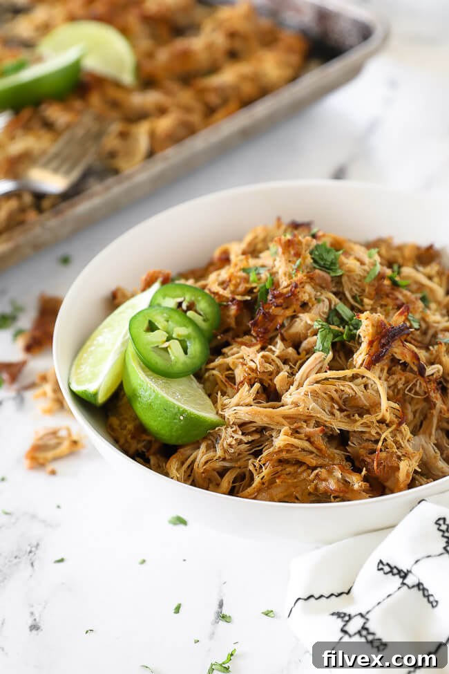 Angled vertical image of chicken carnitas in a bowl with a vibrant garnish of lime wedges, sliced jalapeño, and chopped cilantro, highlighting its fresh appeal.