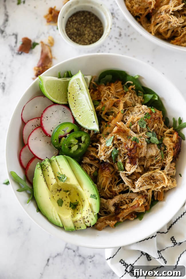 Overhead vertical image of chicken carnitas served in a bowl with fresh greens, thinly sliced radish, bright lemon wedges, vibrant sliced jalapeño, and creamy avocado slices, all topped with fragrant chopped cilantro.