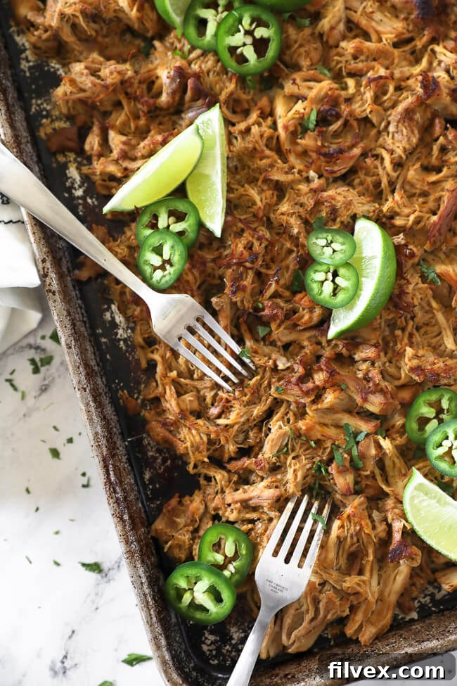 Overhead vertical image of crispy chicken carnitas spread on a sheet pan, ready for shredding, with two forks, fresh lime wedges, sliced jalapeño, and chopped cilantro, indicating preparation for serving.