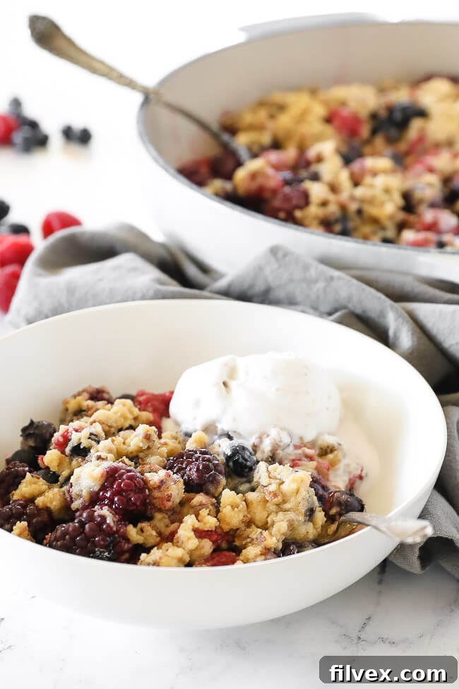 Angled vertical image of berry crisp served in a bowl with a scoop of ice cream on top and a spoon dug in. 