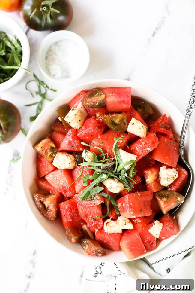 Image of tomato watermelon salad in a bowl with a serving spoon. Fresh basil on top, and flaky sea salt on the side with heirloom tomatoes and more basil. 