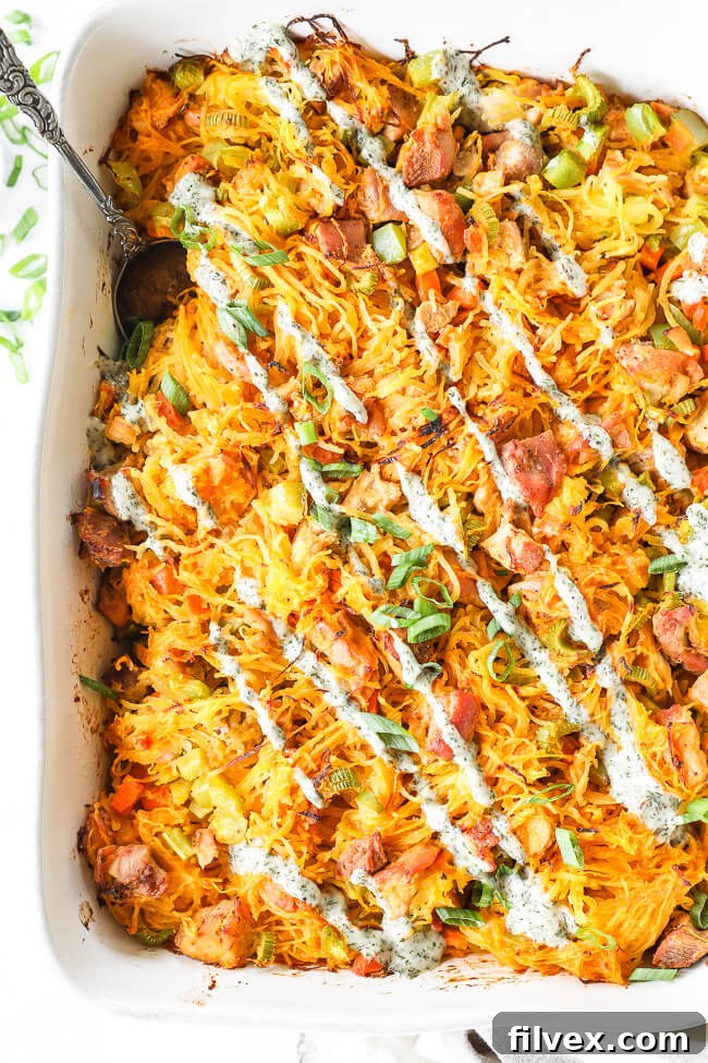 Overhead vertical image of buffalo chicken casserole in casserole dish with chopped green onion and ranch drizzled on top, with a serving spoon ready to dish out a portion.