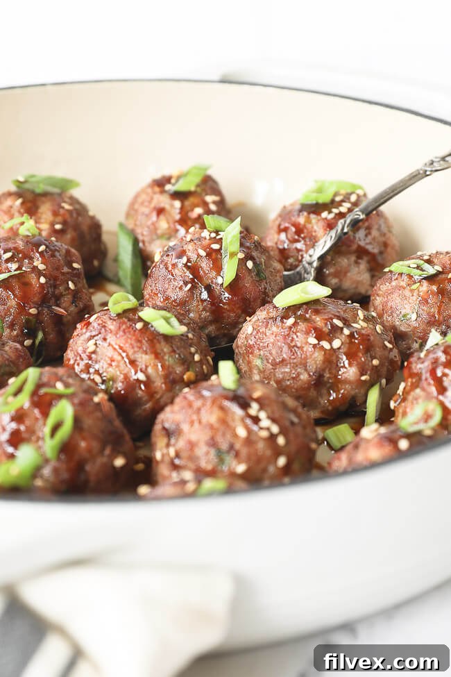 Angled view of perfectly cooked Asian meatballs in a rustic skillet, coated in sauce and sprinkled with fresh green onions and sesame seeds, alongside a serving spoon.