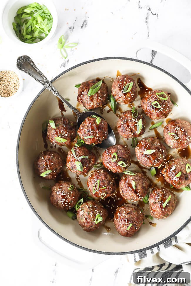 Overhead view of Asian pork meatballs arranged in a skillet, coated in a rich, sticky sauce and topped with fresh green onions and sesame seeds.