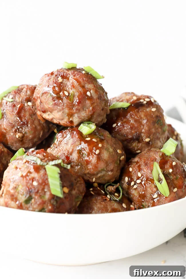 Angled close-up of a bowl filled with Asian pork meatballs, generously coated in sauce, and garnished with fresh green onion and sesame seeds.
