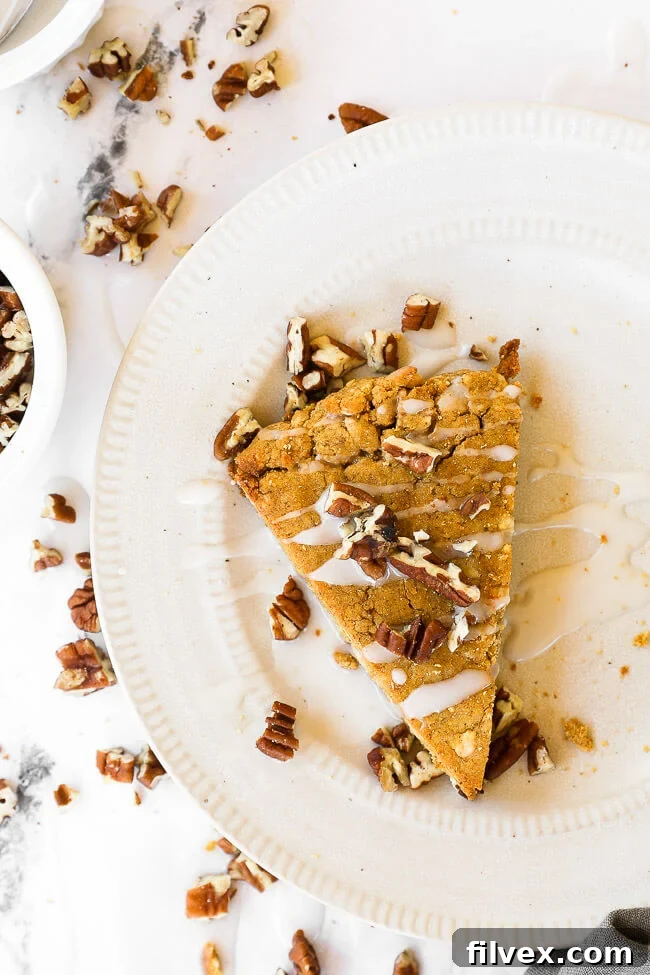 Vertical image of one pumpkin scone on a plate with icing drizzled on top and sprinkled with chopped pecans.