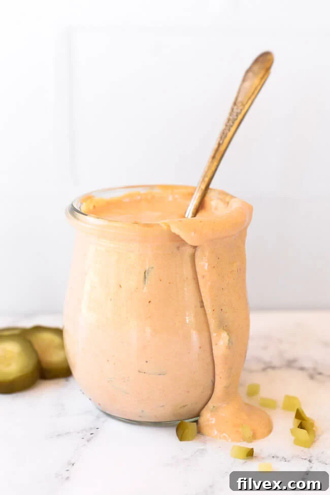 Healthy homemade Big Mac sauce in a clear glass jar with a spoon dipped inside, ready to be served. The sauce is creamy and speckled with finely diced pickles, highlighting its fresh ingredients.