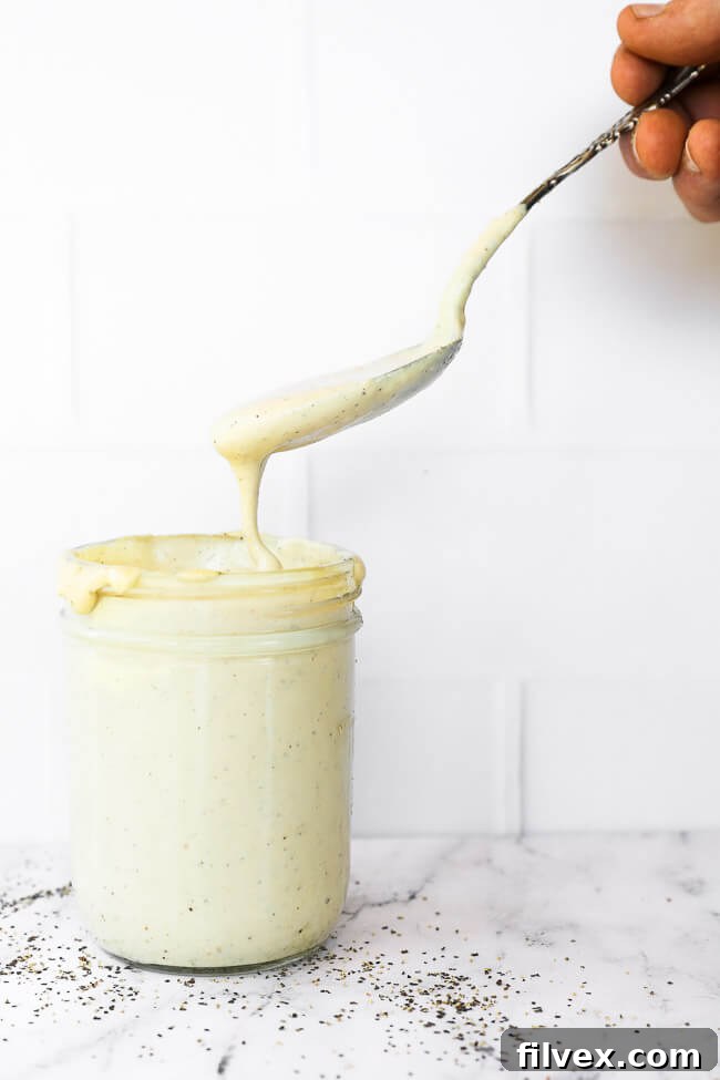 Vertical straight on image of jar of vegan alfredo sauce with a spoonful being pulled out of jar and dripping off spoon. 