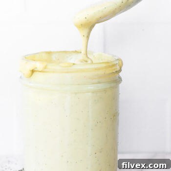 Vertical straight on image of jar of vegan alfredo sauce with a spoonful being pulled out of jar and dripping off spoon.